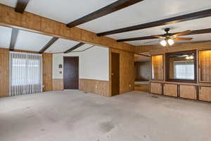 Unfurnished living room featuring wooden walls, carpet flooring, beamed ceiling, a wainscoted wall, and ceiling fan