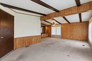 Unfurnished living room with wood walls, carpet flooring, a ceiling fan, wainscoting, and lofted ceiling with beams