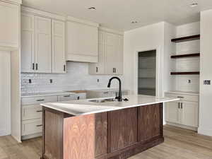 Two tone kitchen featuring open shelves, backsplash, dual tone cabinetry, light wood-type flooring, and a center island with sink