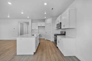 Kitchen featuring white cabinetry, stainless steel appliances, a center island with sink, light wood finished floors, and light stone counters