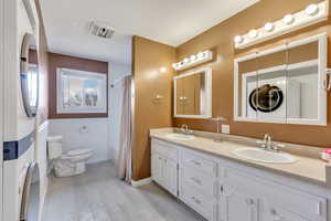 Bathroom featuring shower / bathtub combination with curtain, double vanity, wainscoting, stacked washer / dryer, and light wood-type flooring
