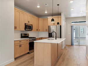 Kitchen featuring stainless steel appliances, light wood finish cabinetry, pendant lighting, a center island with sink, and light wood-type flooring