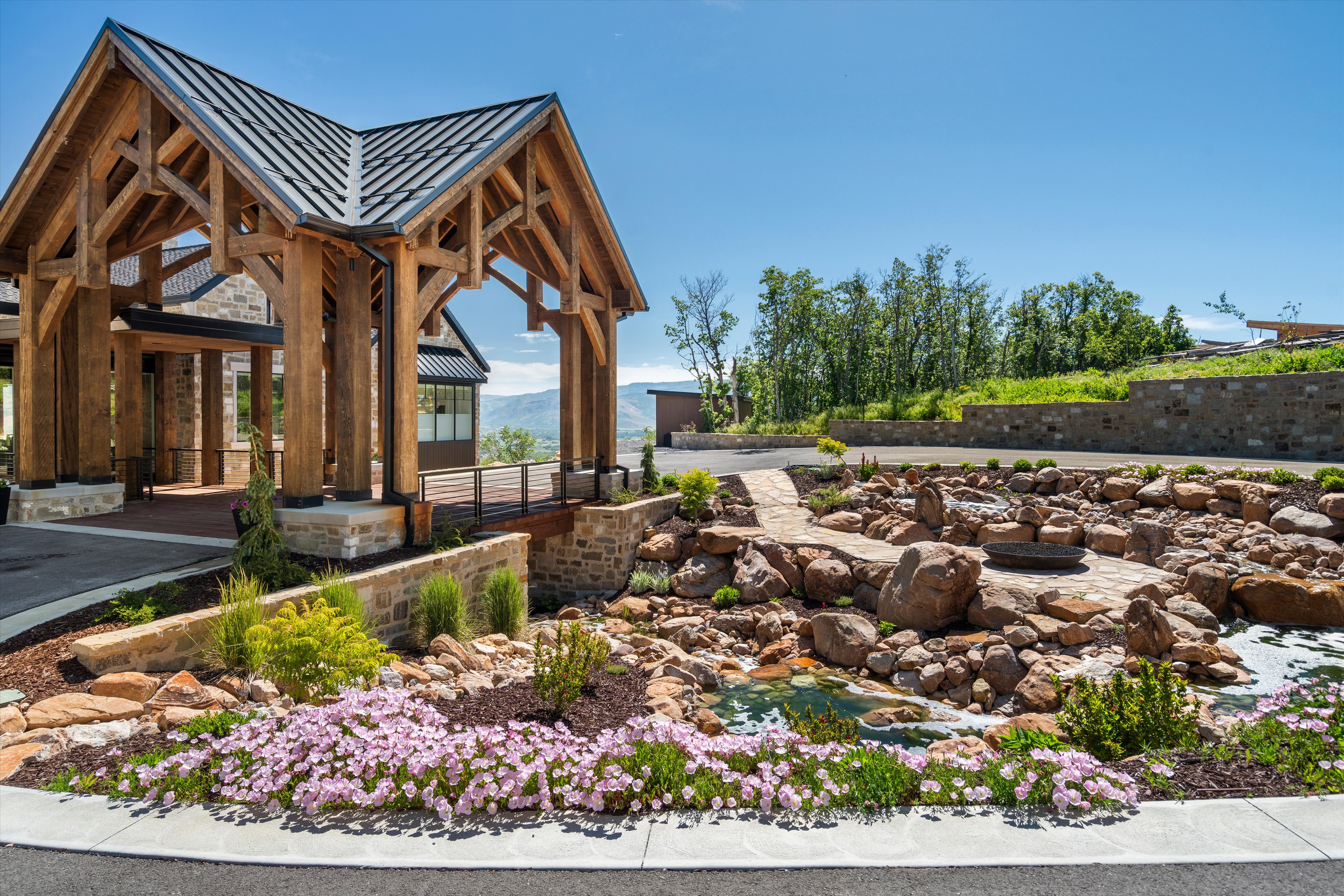 Timber-framed porte-cochère with a metal roof, stone accents, and a naturalistic rock garden featuring a pond and integrated fire pit area