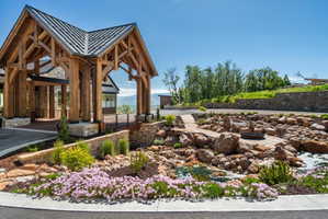 Timber-framed porte-cochère with a metal roof, stone accents, and a naturalistic rock garden featuring a pond and integrated fire pit area