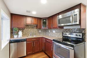 Kitchen with stainless steel appliances, light stone counters, light wood finished floors, glass fronted cabinets, and tasteful backsplash