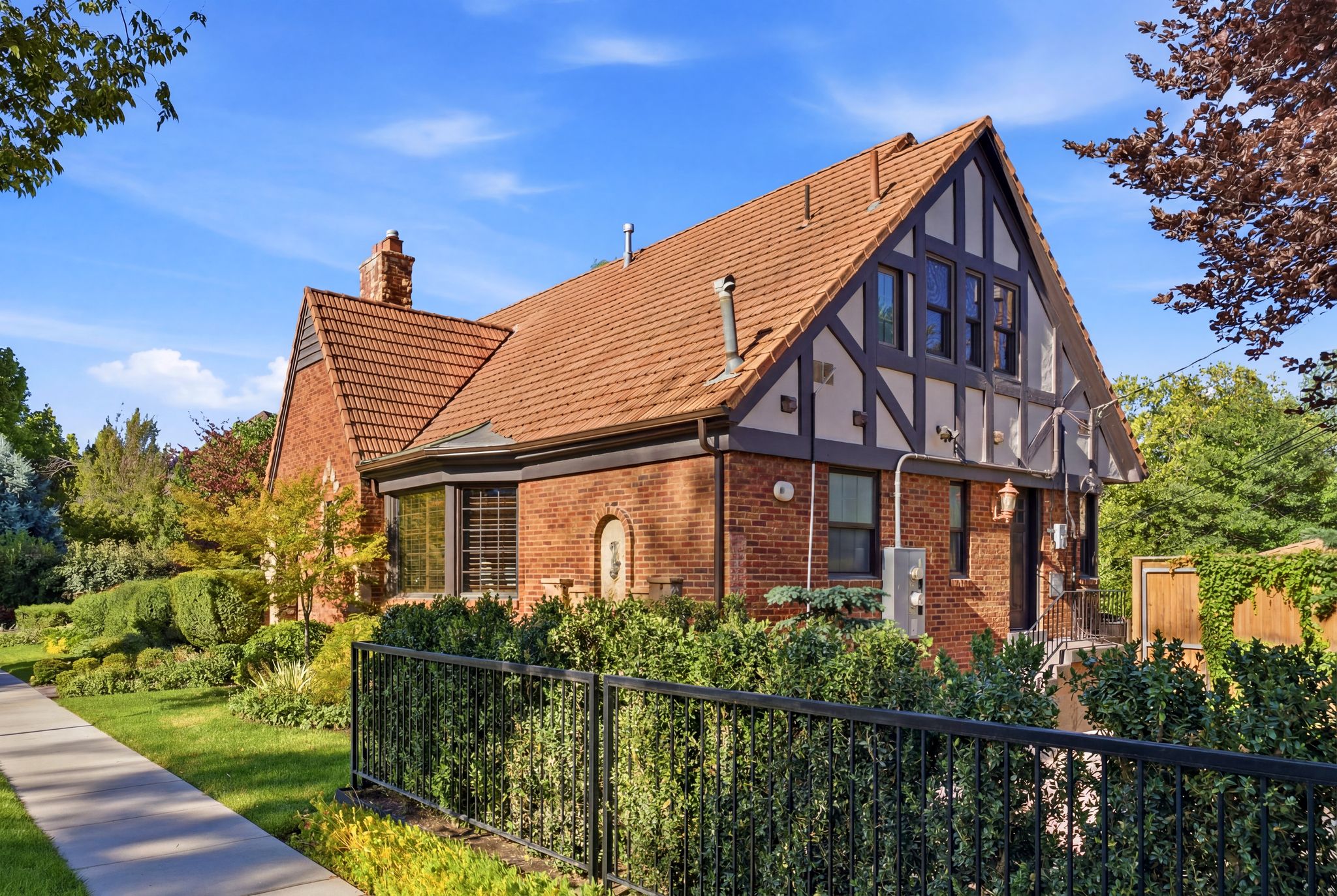 Brick and half-timbered exterior featuring a terracotta tile roof, prominent chimney, bay window, and a wrought iron fence bordering manicured hedges