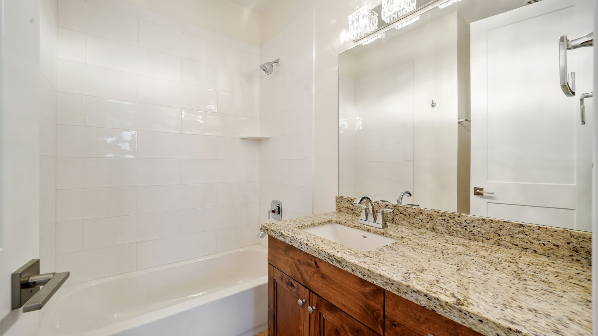 Bathroom with a white tiled shower/tub combination, a wood-finish vanity, a light-toned stone countertop, and a large mirror with a multi-bulb light fixture