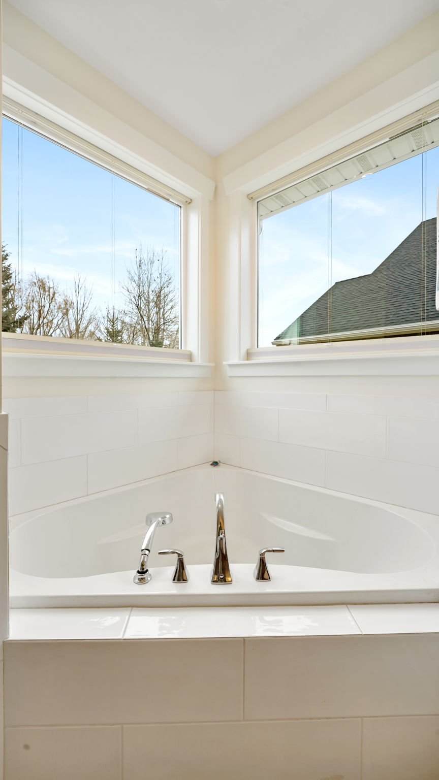 Corner soaking tub with polished chrome fixtures, surrounded by white subway tile and two large windows