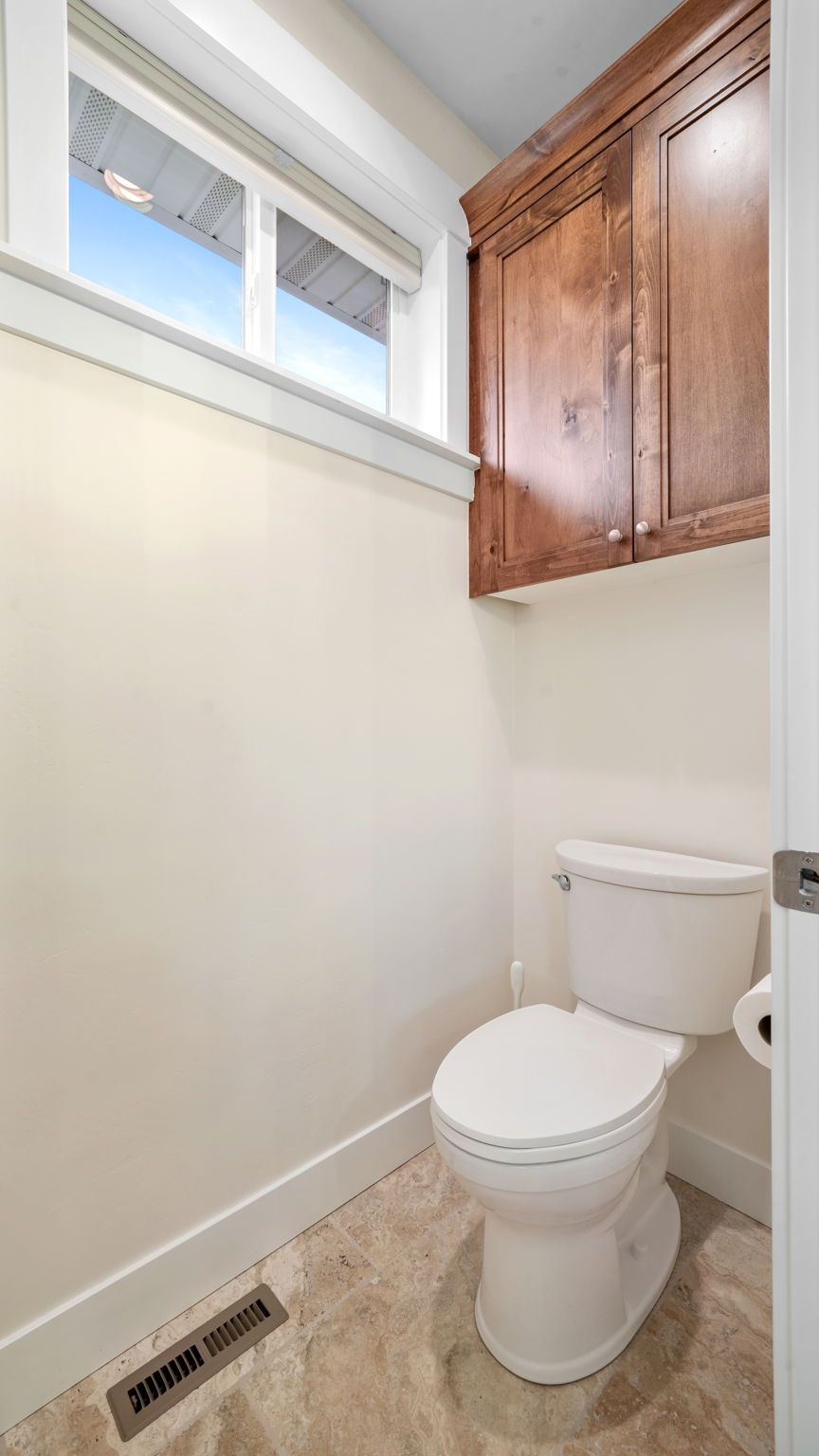 Powder room featuring a white toilet, wood-finish wall-mounted cabinetry, a window with white trim, and tiled flooring