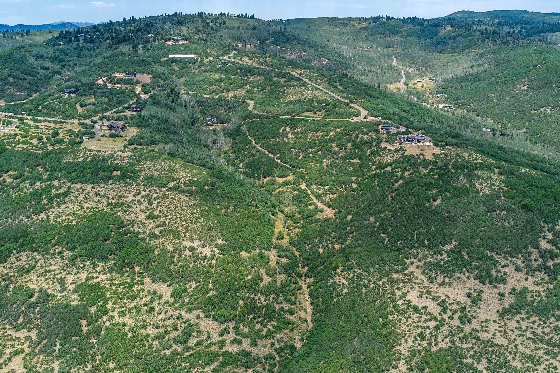 Expansive mountainous terrain featuring scattered residential structures, abundant green vegetation, natural pathways, and distant tree lines