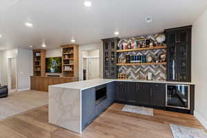 Indoor dry bar featuring open shelves, light stone counters, wine cooler, glass fronted cabinets, and stainless steel microwave