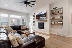 Living area featuring built in shelves, light wood finished floors, ceiling fan, a glass covered fireplace, and a textured ceiling