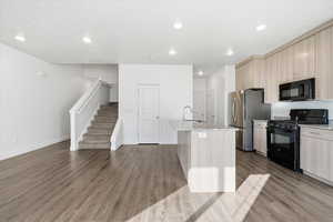 Kitchen with black appliances, light stone counters, a kitchen island with sink, light wood finish cabinets, and dark wood-style floors