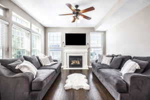Living room featuring dark wood-type flooring, a ceiling fan, a fireplace with flush hearth, and plenty of natural light