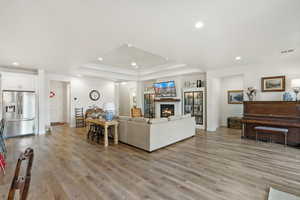 Living room with ceiling fan, a warm lit fireplace, light wood finished floors, recessed lighting, and a raised ceiling