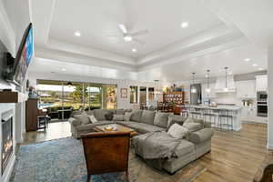 Living area featuring a ceiling fan, recessed lighting, light wood-style floors, a glass covered fireplace, and a tray ceiling