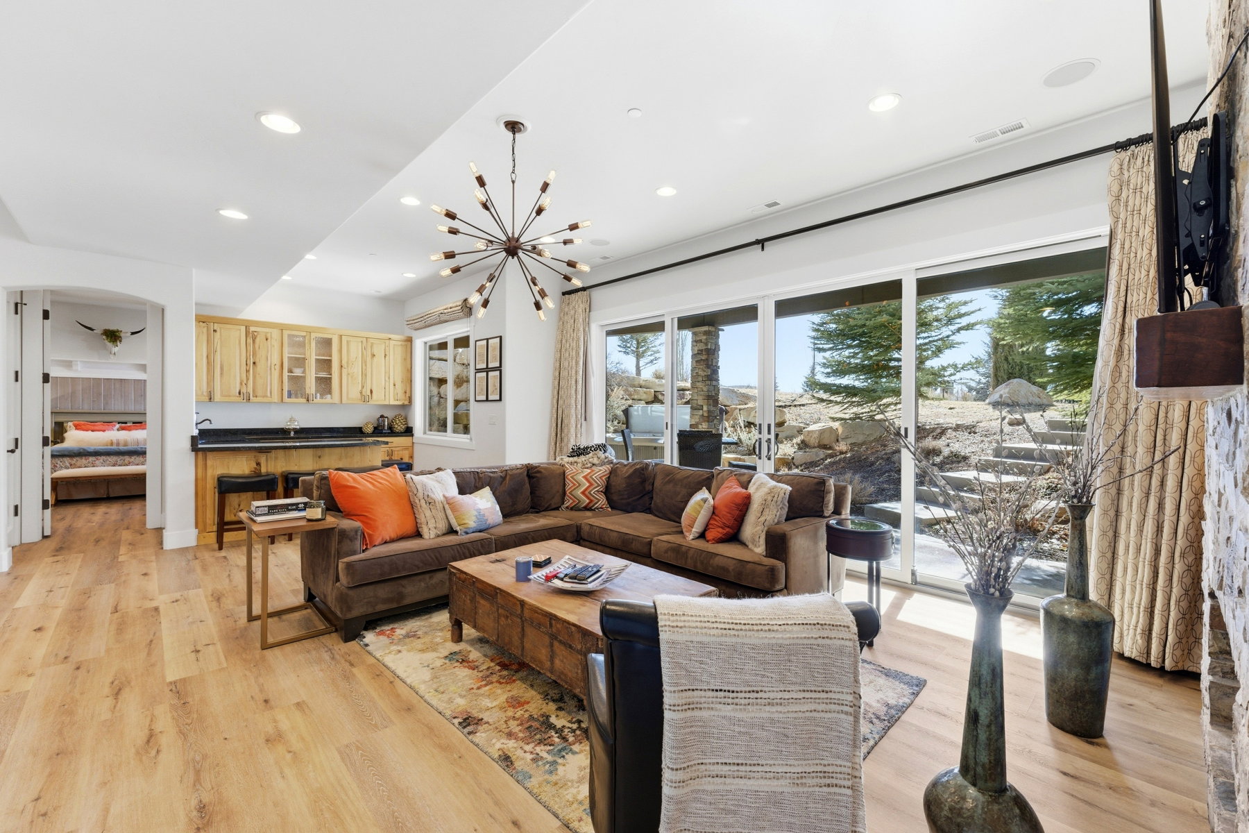 Spacious living area featuring light wood-finish flooring, a contemporary sputnik chandelier, and a built-in wet bar with wood cabinetry and a dark countertop