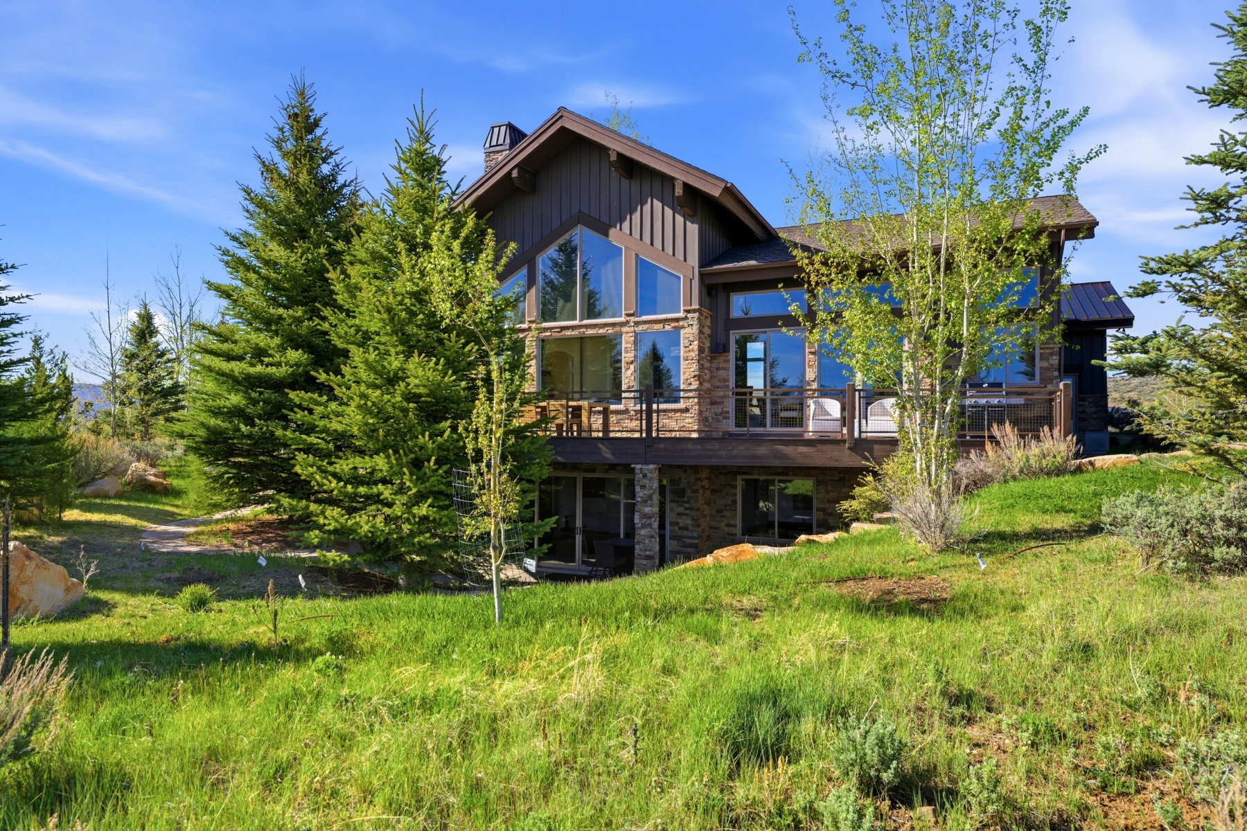 Mountain-style residence featuring extensive window walls, a prominent stone facade, wood siding, multiple outdoor decks, and a sloping natural landscape