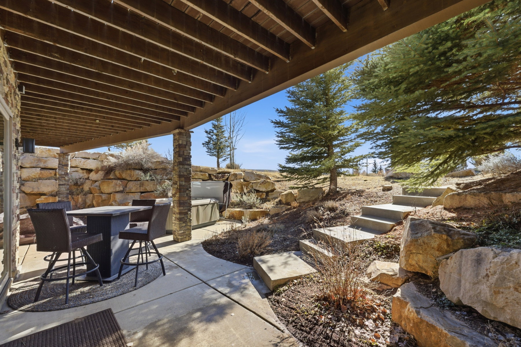 Covered concrete patio featuring exposed wood beam ceiling, stone-clad support columns, and built-in hot tub