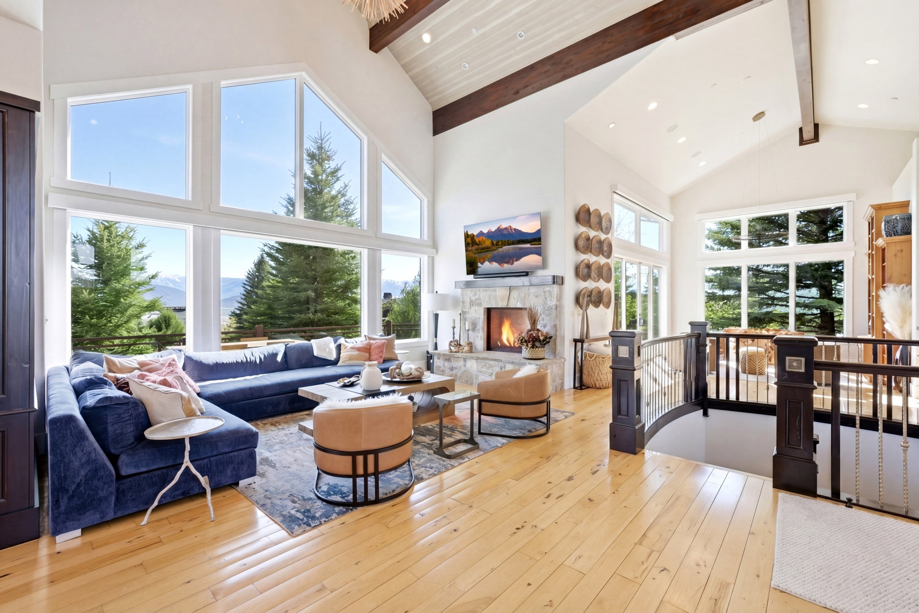 Vaulted ceilings featuring exposed wood beams, a stone fireplace, light wood-finish flooring, and a railing with dark wood newel posts and metal spindles