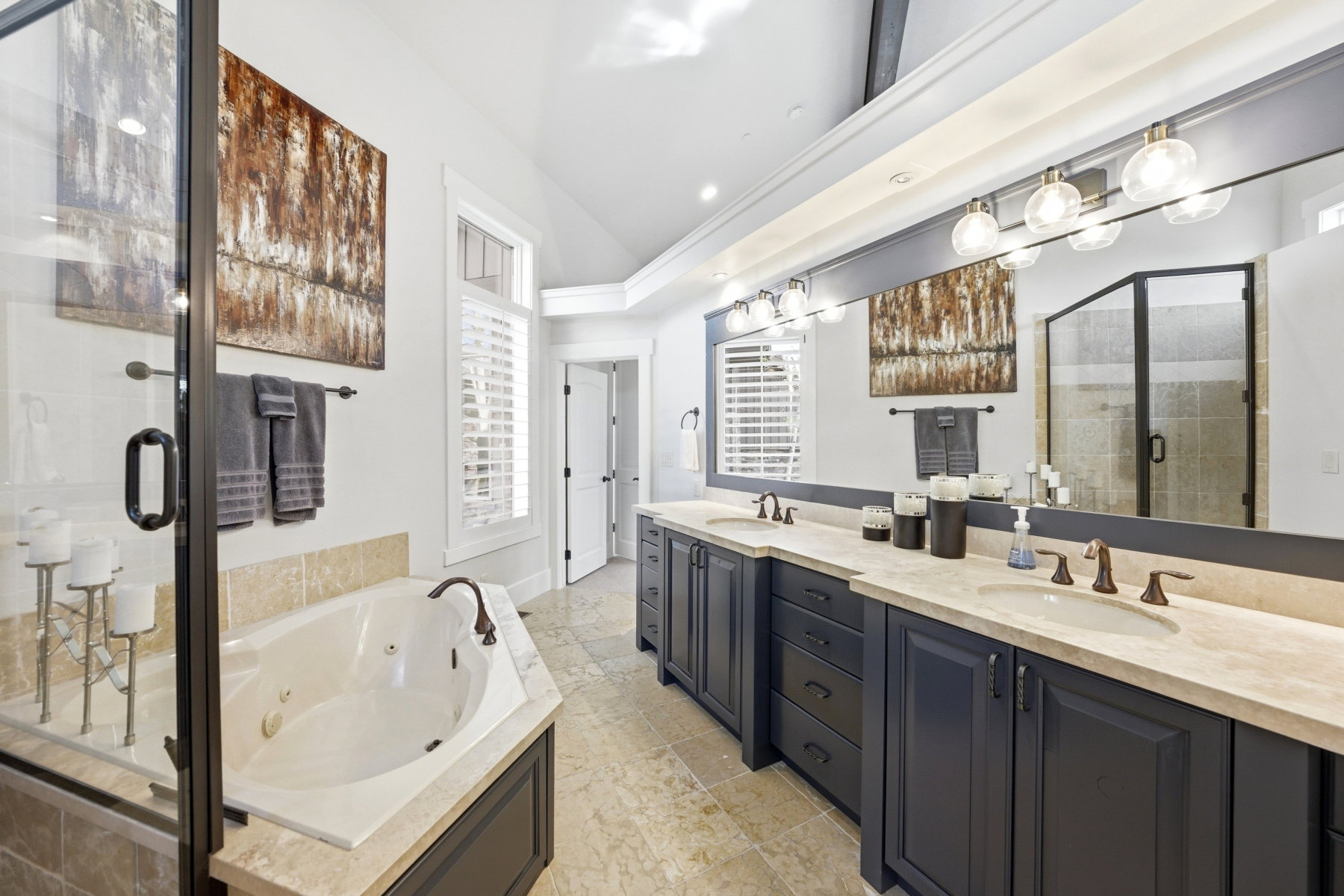 Spacious bathroom featuring a built-in jetted tub with tile surround, a large walk-in shower with glass enclosure, dual vanities with undermount sinks and bronze-finish fixtures, and plantation shutters on windows