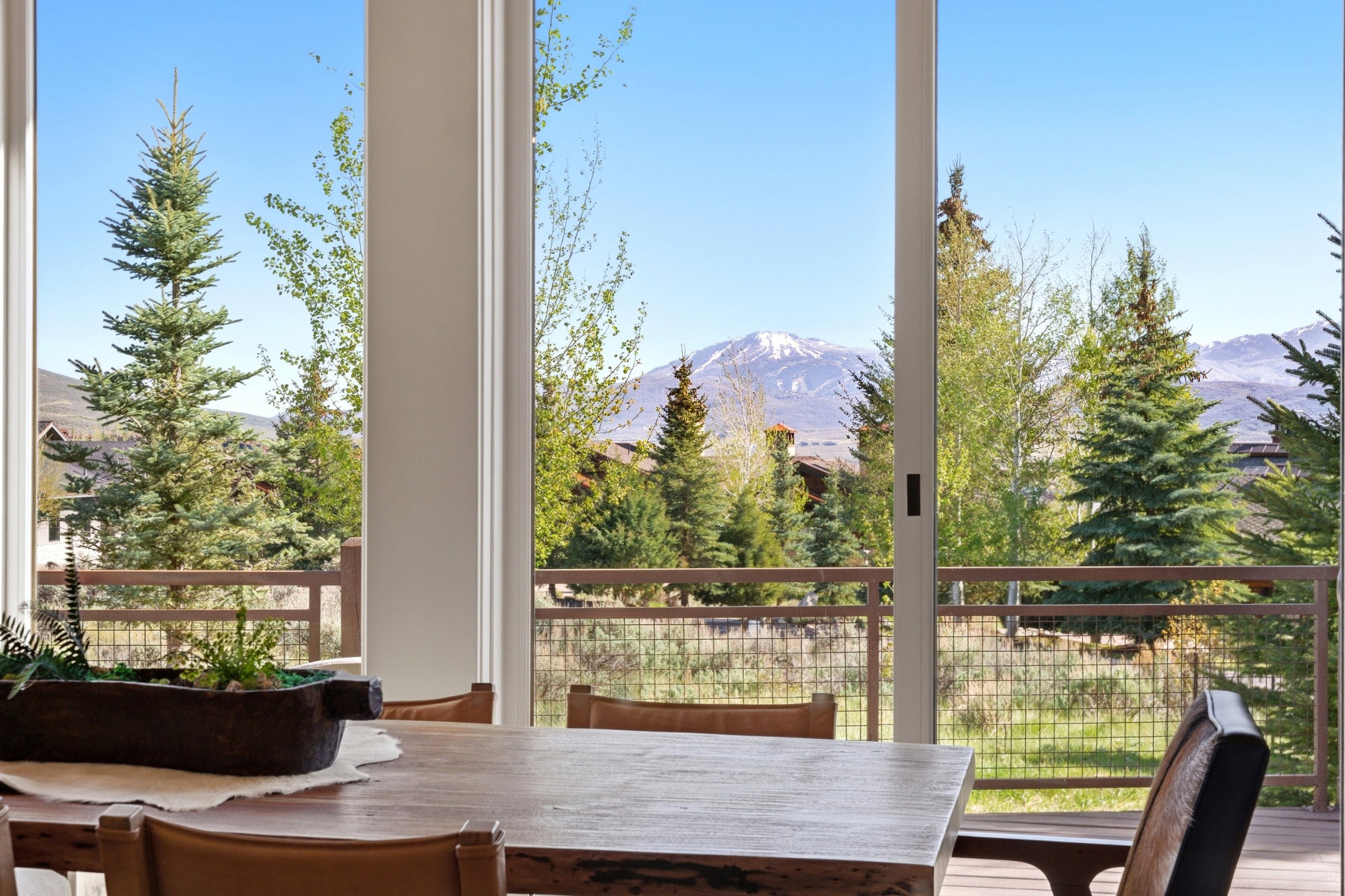 Expansive glass sliding doors open to an outdoor deck with a metal and wood railing, offering mountain scenery and surrounding mature trees