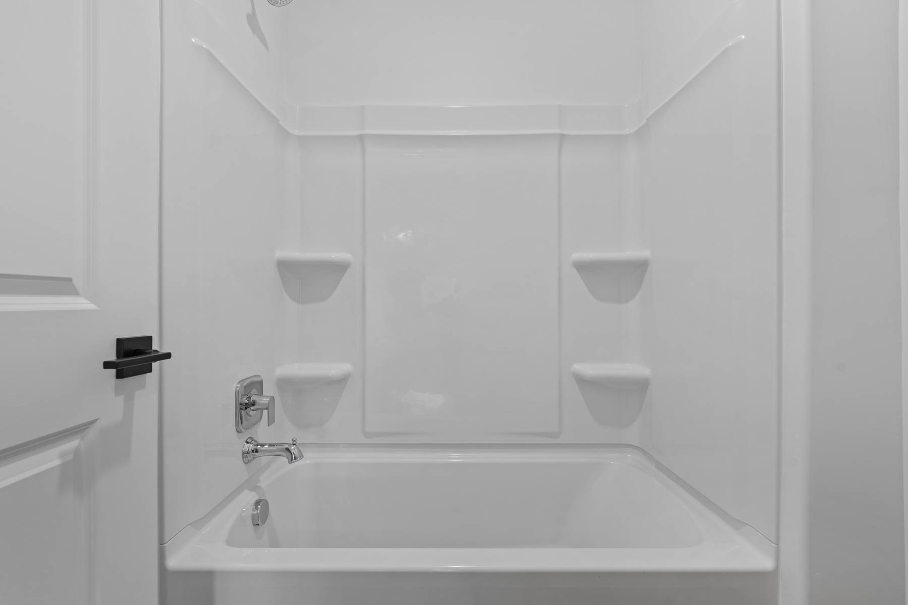 White fiberglass tub and shower combination with integrated shelving, chrome fixtures, and a matte black door handle