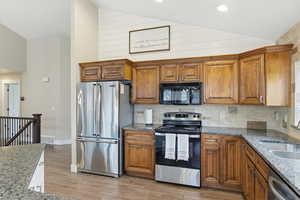 Kitchen featuring lofted ceiling, dark stone counters, stainless steel appliances, wood finish cabinets, and light wood-style floors