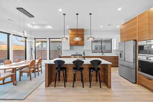 Kitchen with modern cabinets, wood finish cabinetry, pendant lighting, stainless steel appliances, and a kitchen bar