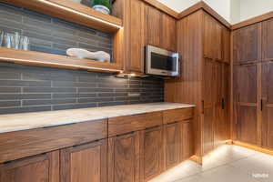 Kitchen featuring wood finish cabinetry, backsplash, open shelves, stainless steel microwave, and light stone counters