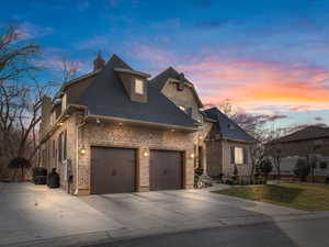 French country inspired facade with brick siding, driveway, a shingled roof, a yard, and a garage