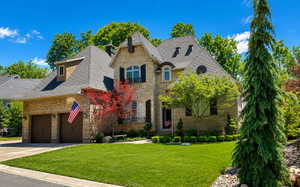 Stone and brick exterior residence featuring a multi-gabled roofline with dormers, a two-car garage with dark wood-look doors, and a manicured front lawn
