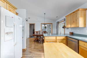 Kitchen with white fridge with ice dispenser, vaulted ceiling, stainless steel dishwasher, light wood finish cabinetry, and tasteful backsplash