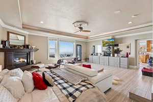 Bedroom with a tray ceiling, a glass covered fireplace, wood finished floors, crown molding, and a ceiling fan