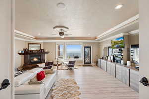 Living room with a tray ceiling, a glass covered fireplace, a ceiling fan, light wood-type flooring, and a textured ceiling