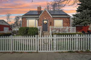 Bungalow-style house featuring brick siding, a chimney, roof mounted solar panels, a fenced front yard, and a gate