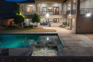 Back of house at twilight featuring a patio, a balcony, stucco siding, a pool with connected hot tub, and a ceiling fan