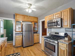 Kitchen with stainless steel appliances, ceiling fan, glass insert cabinets, light wood-type flooring, and light stone countertops