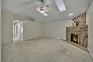 Unfurnished living room featuring a fireplace, ceiling fan, vaulted ceiling, light carpet, and a skylight