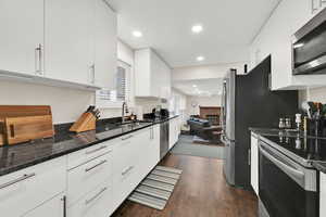 Kitchen with a brick fireplace, dark stone countertops, stainless steel appliances, white cabinetry, and dark wood finished floors