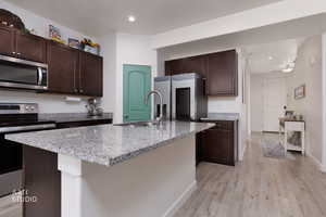 Kitchen with dark wood finish cabinetry, stainless steel appliances, light wood finished floors, light stone countertops, and a kitchen island with sink