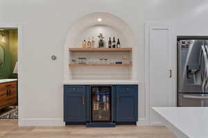 Indoor dry bar with open shelves, stainless steel refrigerator with ice dispenser, wine cooler, light stone counters, and blue cabinetry