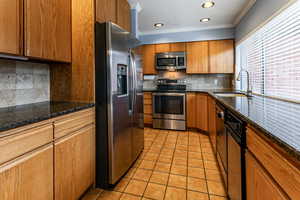 Kitchen with stainless steel appliances, crown molding, dark stone countertops, wood finish cabinets, and tasteful backsplash