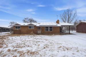 Snow covered rear of property with roof mounted solar panels, a chimney, brick siding, an attached carport, and a patio area