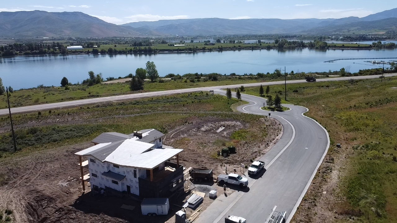 Expansive land parcel featuring an unfinished residential structure, a paved winding road, and a substantial body of water with distant mountains