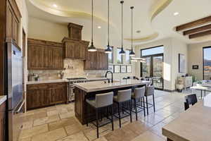 Kitchen featuring a breakfast bar, a raised ceiling, an island with sink, high end appliances, and stone tile floors