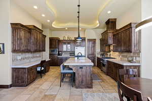 Kitchen featuring dark wood finish cabinetry, a tray ceiling, built in appliances, stone tile floors, and a kitchen island with sink
