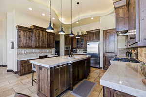 Kitchen with dark wood finish cabinetry, a kitchen island with sink, built in appliances, stone tile flooring, and glass fronted cabinets