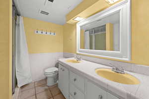 Bathroom with a shower stall, double vanity, tile walls, a wainscoted wall, and light tile patterned floors