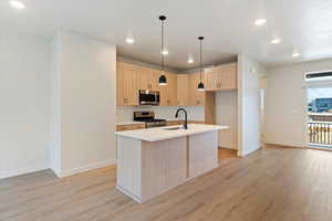Kitchen with light wood finish cabinetry, light wood-type flooring, stainless steel appliances, hanging light fixtures, and a kitchen island with sink
